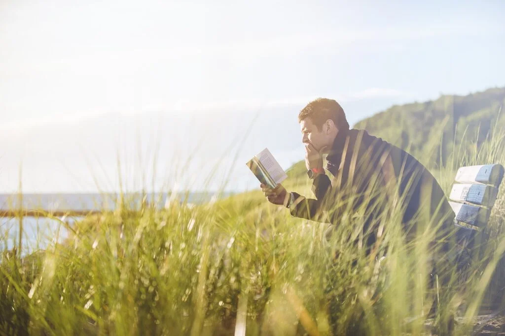 man reading on a bench on a hill next to a body of water