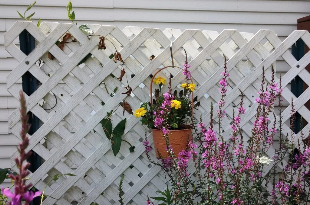 An orange basket of yellow flowers on a white fence with pink flowers next to it.